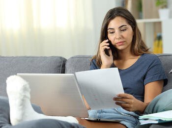 Frau telefoniert mit Gipsfuß, Laptop und Zettel auf Sofa