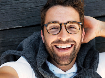 Ein Mann mit Bart und kurzen braunen Haaren träft eine braun gemusterte Brille und lacht ausgelassen in die Kamera.