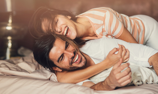 Eine junge Frau umarmt einen jungen Mann. Sie liegen im Bett und lachen ausgelassen.