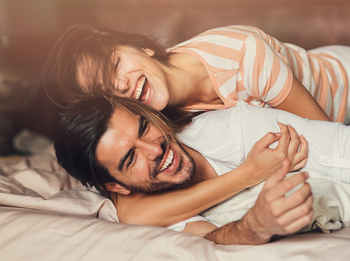 Eine junge Frau umarmt einen jungen Mann. Sie liegen im Bett und lachen ausgelassen.
