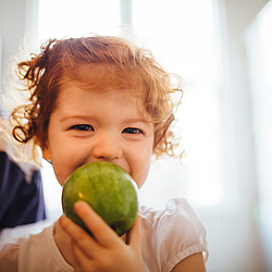 Ein kleines Mädchen mit roten lockigen Haaren beißt in einen großen grünen Apfel. Sie lacht dabei in die Kamera. 