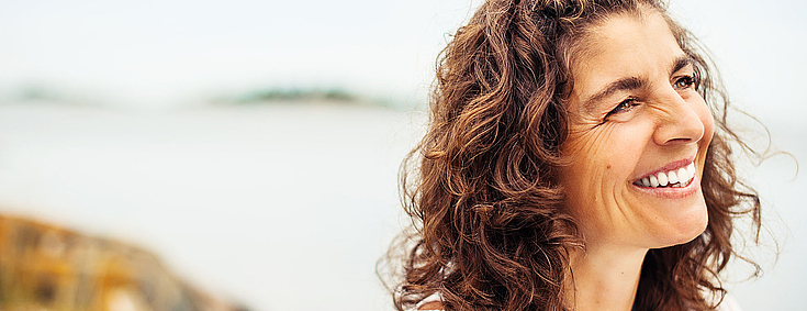 Frau mittleren Alters, dunkelbraune lockige Haare, schaut an dem Betrachter vorbei. zur rechten Bildseite, sie lacht und sieht glücklich aus. Im Hintergrund kann man einen Strand und das Meer erahnen. 