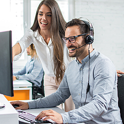 Ein Mann sitzt lächelnd vor einem Monitor. Er trägt ein Headset. Neben ihm steht eine Frau die ihm etwas auf dem Monitor zeigt.