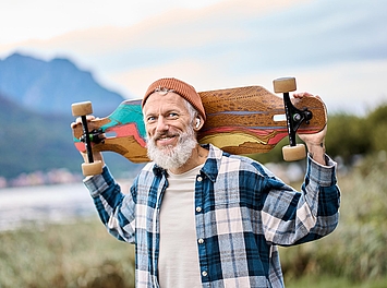 Lächelnde Person mit grauem Bart trägt ein Longboard über den Schultern und blickt in die Kamera.