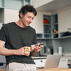 Ein Mann steht in seiner Küche und schaut auf sein Smartphone. In der anderen Hand hält er eine Tasse.