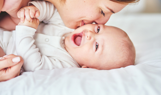 Ein Baby in einem weißen Body liegt auf einer Matratze. Es lacht fröhlich in die Kamera. Seine Mutter lächelt und gibt ihm einen Kuss auf die Wange.