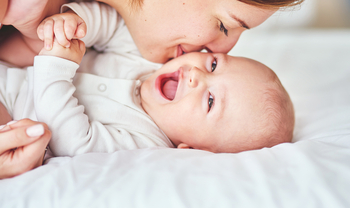 Ein Baby in einem weißen Body liegt auf einer Matratze. Es lacht fröhlich in die Kamera. Seine Mutter lächelt und gibt ihm einen Kuss auf die Wange.