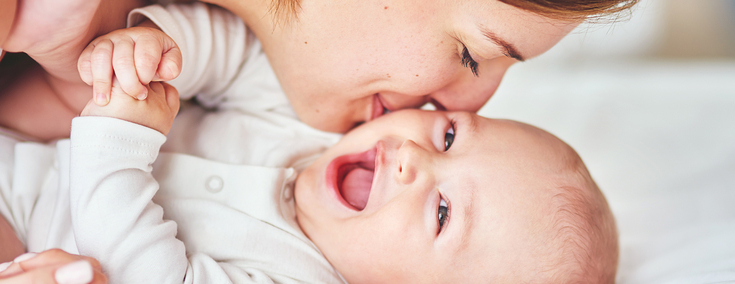 Ein Baby in einem weißen Body liegt auf einer Matratze. Es lacht fröhlich in die Kamera. Seine Mutter lächelt und gibt ihm einen Kuss auf die Wange.