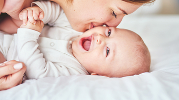 Ein Baby in einem weißen Body liegt auf einer Matratze. Es lacht fröhlich in die Kamera. Seine Mutter lächelt und gibt ihm einen Kuss auf die Wange.