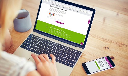 Eine junge Frau sitzt an einem Laptop. Auf dem Bildschirm sieht man die Internetseite der BKK GS. Auf dem Tisch liegt ihr Smartphone mit der BKK GS App.