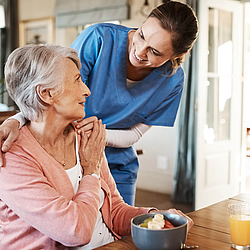 Eine ältere Frau sitzt am Tisch. Ihre Pflegerin legt einen Arm um sie und beide lächeln sich an.