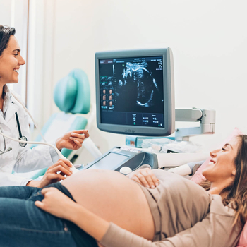 Eine junge schwangere Frau liegt auf einer Untersuchungsliege. Sie lächelt eine junge Ärtzin an, die einen Ultraschall von ihrem Baby macht.