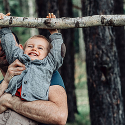 Ein Vater hebt seinen Sohn im Wald hoch, damit der Sohn an einem Ast hängen kann. Sie lachen beide dabei.