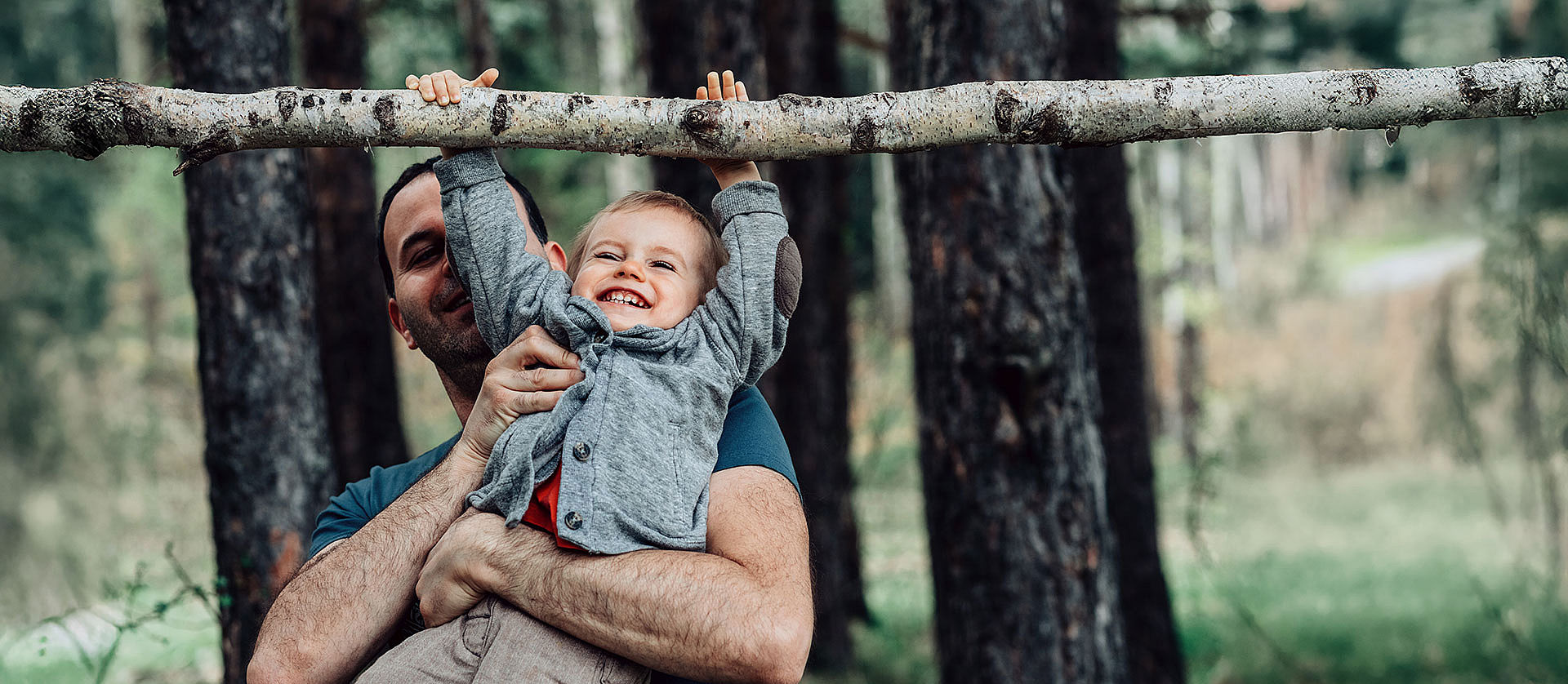 Ein Vater hebt seinen Sohn im Wald hoch, damit der Sohn an einem Ast hängen kann. Sie lachen beide dabei.