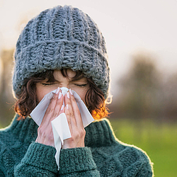 Eine junge Frau mit grauer Wollmütze schnäuzt in ein Taschentuch.