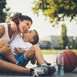 Eine Mutter sitzt mit ihrem kleinen Sohn draußen auf dem Boden. Er hat einen Arm um sie gelegt. Neben ihnen liegt ein Basketball.