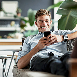 Ein junger Mann sitzt entspannt auf dem Sofa und schaut auf sein Smartphone.