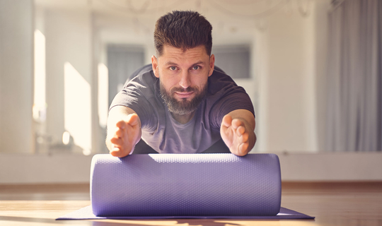 Ein Mann mit Bart sitzt auf einer Trainingsmatte und macht Übungen mit einer Faszienrolle.