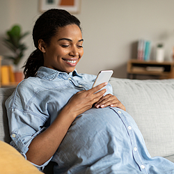 Eine schwangere Frau sitzt auf einem Sofa und schaut auf ihr Smartphone.