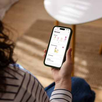 Eine Frau mit lockigen Haaren sitzt in ihrem Wohnzimmer. Auf ihrem Smartphone nutzt sie die ePA App.