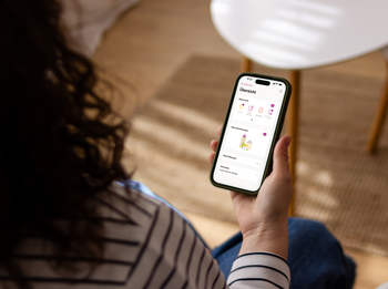 Eine Frau mit lockigen Haaren sitzt in ihrem Wohnzimmer. Auf ihrem Smartphone nutzt sie die ePA App.