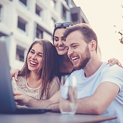 Eine Gruppe junger Menschen sitzen im Außenbereich eines Restaurants. Sie schauen gemeinsam auf einen Laptop und lachen.