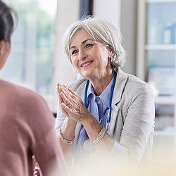 Eine ältere Hausärztin mit kurzen grauen Haaren führt ein Arztgespräch mit einer Patientin. Sie lächelt dabei freundlich.