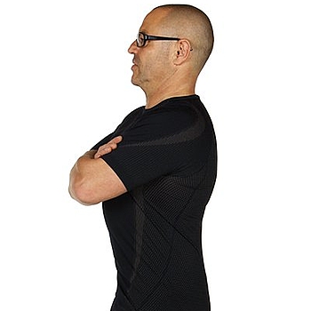 Ein Mann mit Brille trägt schwarze Kleidung. Er steht gerade und hat die Arme überkreuzt. Er zeigt die Übung Beckenwippe.