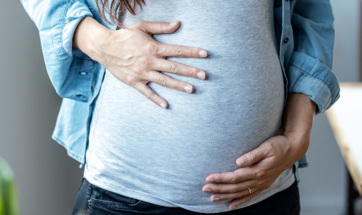 Eine junge Frau trägt ein graues Shirt und ein Jeanshemd. Sie hat die Hände oben und unten auf ihren schwangeren Bauch gelegt.