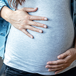 Eine Frau trägt ein graues Shirt und ein Jeanshemd. Sie hat die Hände oben und unten auf ihren schwangeren Bauch gelegt.