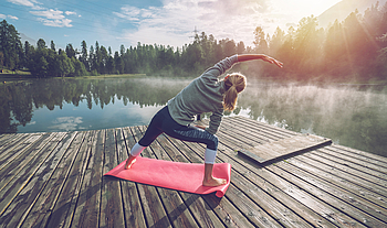 Eine Frau macht Yoga-Übungen an einem See. Sie steht dabei auf einem Steg.