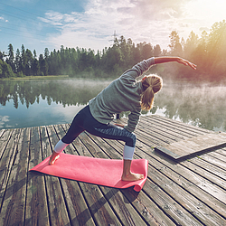 Eine Frau macht Yoga-Übungen an einem See. Sie steht dabei auf einem Steg.