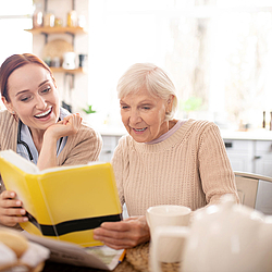 Eine junge Pflegerin sitzt mit einer älteren Frau am Küchentisch. Gemeinsam schauen sie sich ein Buch an und lachen.