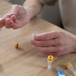 Eine Person führt einen medizinischen Selbsttest durch. Dazu hat hat sie sich in den Finger gestochen und sammelt das Blut in einem Probenröhrchen.