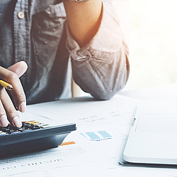 Eine Person sitzt am Tisch, zwischen den Finger hält sie einen gelben Bleistift und gibt etwas in einem Taschenrechner ein.