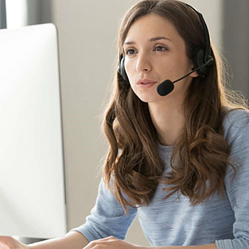 Eine junge Frau mit langen braunen Haaren trägt ein hellblaues Oberteil. Sie sitzt vor einem großen weißen Bildschirm und trägt ein Headset. Sie spricht in das Mikrofon, während sie auf den Bildschirm schaut.