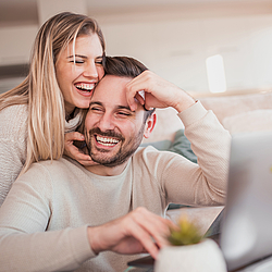 Ein junges Pärchen sitzt auf dem Sofa und lächelt.