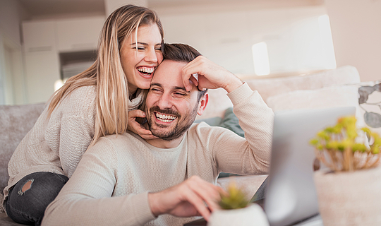 Ein junges Pärchen sitzt auf dem Sofa und lächelt.