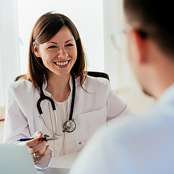 Eine junge Ärtzin mit schulterlangen braunen Haaren lacht ihren Patienten über den Tisch hinweg an.