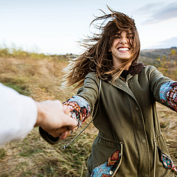 Eine Frau mit braunen Haaren hält von jemandem die Hände. Sie lächelt und wird von der anderen Person umhergewirbelt. Ihre Haare wehen im Wind.