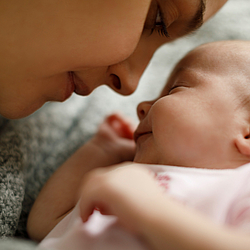 Eine junge Mutter beugt sich nah zu ihrem Baby, sie lächelt. Das Baby hat die Augen geschlossen. 