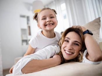 Eine junge Frau mit langen blonden Haaren liegt auf dem Bauch auf ihrem beigen Sofa. Auf ihrem Rücken liegt ihre kleine Tochter. Beide lachen ausgelassen in die Kamera.