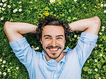 Ein Mann mit braunen Locken liegt entspannt auf eine Wiese und lächelt.