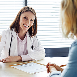 Eine lächelnde Ärztin mit schulterlangen braunen Haaren sitzt mit einer Patientin am Tisch. Sie unterhalten sich.