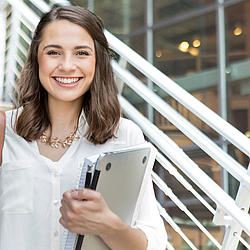 Eine Frau steht lächelnd vor einer Treppe. Sie hält einen Notizblock und Laptop in der Hand.