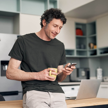 Ein Mann mit lockigen Haaren steht in einer Küche und hält eine gelbe Tasse in der Hand. Mit der anderen Hand bedient er sein Handy.