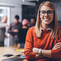 Eine Frau mit Brille schaut lächelt. Sie trägt einen roten Pullover. Im Hintergrund stehen mehrere Personen zusammen.
