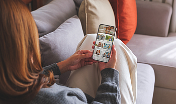 Eine Frau mit roten langen Haaren sitzt auf dem Sofa. Sie benutzt die BKK GS-App auf ihrem Smartphone. 
