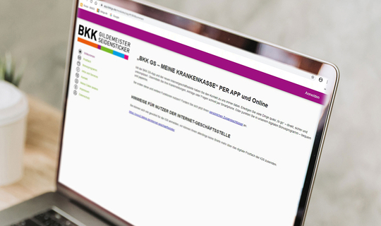 Eine junge Frau sitzt vor einem Laptop. Auf dem Bildschirm ist die Internetseite der BKK GS. In ihrer linken Hand hält sie ein Smartphone mit der BKK GS App und in ihrer linken Hand die BKK Versichertenkarte. 