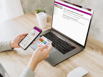Eine junge Frau sitzt vor einem Laptop. Auf dem Bildschirm ist die Internetseite der BKK GS. In ihrer linken Hand hält sie ein Smartphone mit der BKK GS App und in ihrer linken Hand die BKK Versichertenkarte. 